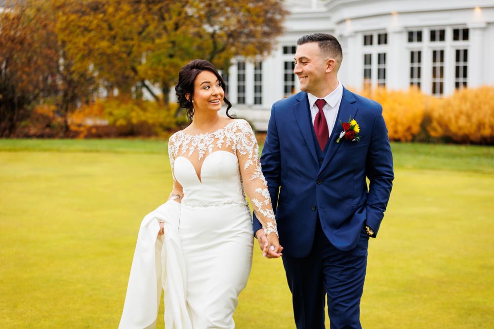 Bride and groom holding hands walking on green lawn with autumn foliage backdrop.