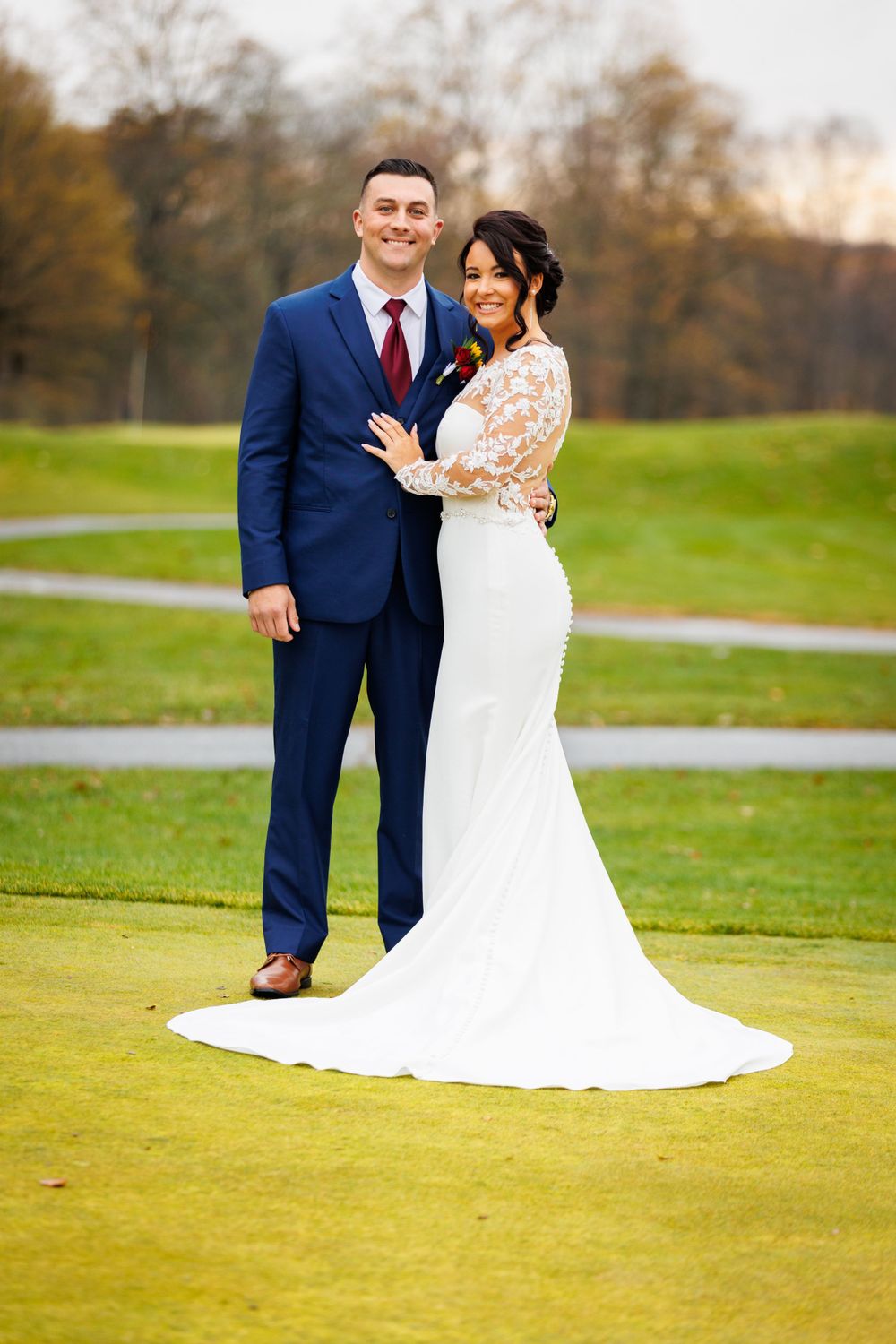 Bride in lace gown and groom in navy suit posing on golf course in autumn.