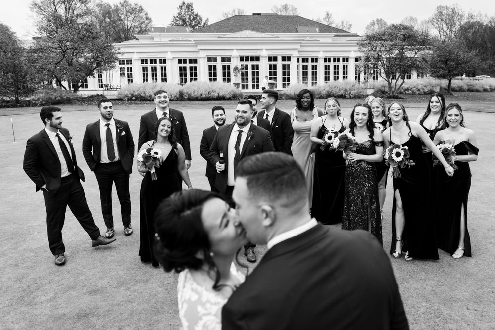 Bride and groom share a kiss while wedding party poses behind them on a golf course in black and white.