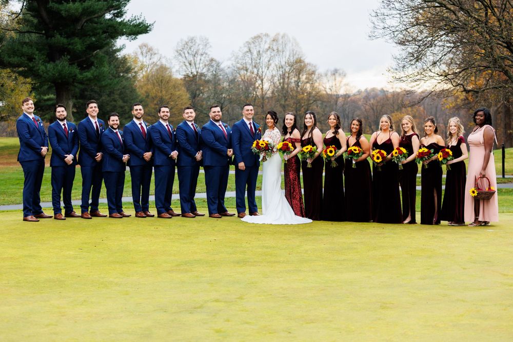 Wedding party poses on golf course, groomsmen in navy suits and bridesmaids in dark gowns with sunflowers.