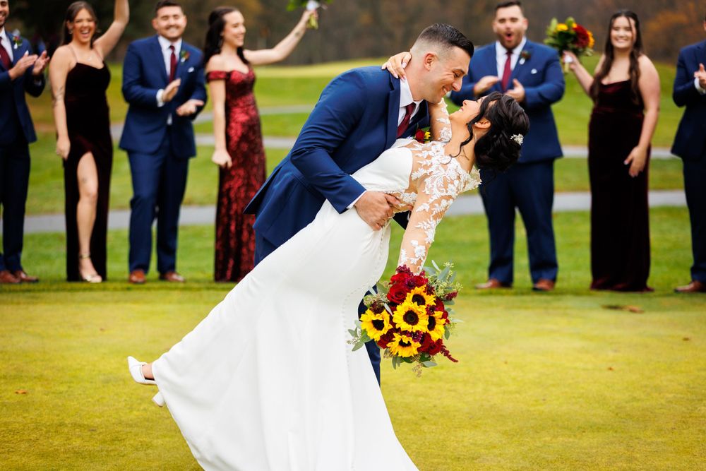 Groom dips bride holding sunflower bouquet while wedding party celebrates behind them on a green lawn.