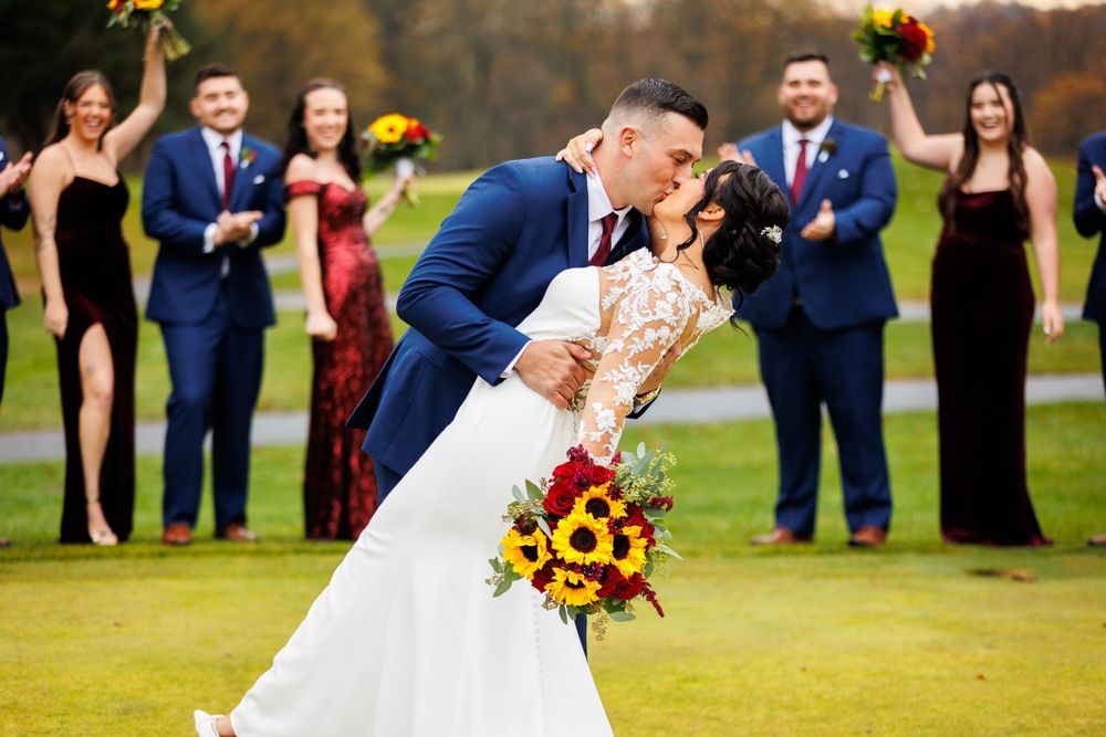 Groom kisses bride in lace gown holding sunflower bouquet as wedding party cheers on autumn golf course.