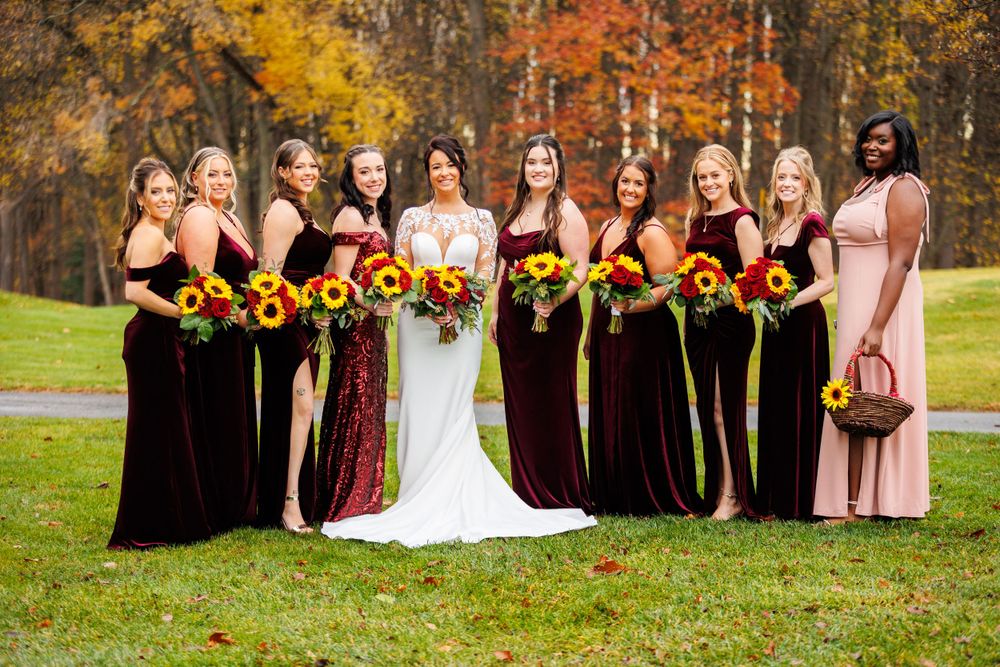 Bride with bridesmaids in dark burgundy gowns holding vibrant sunflower bouquets on an autumn golf course.