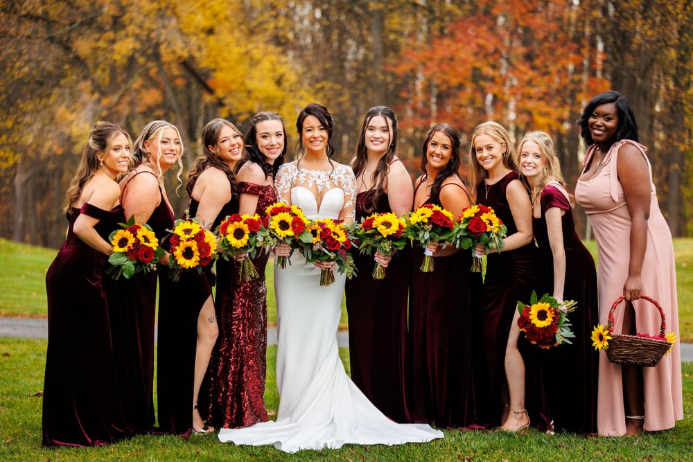 Bride and bridesmaids in burgundy velvet gowns holding sunflower bouquets pose before colorful fall foliage.