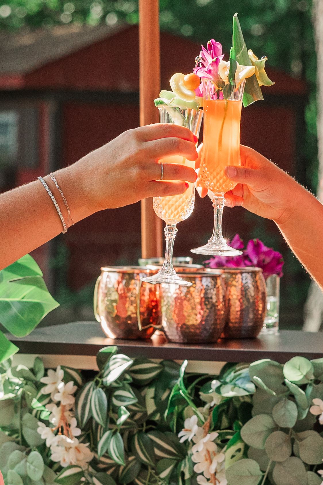 Two hands toasting orange cocktails decorated with flowers against a leafy backdrop and red building.