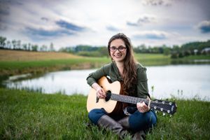 Woman plays acoustic guitar while sitting by lake on sunny day.