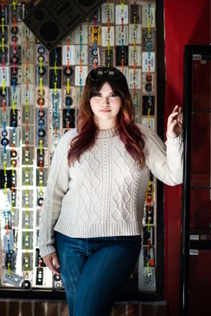 Woman in cream cable knit sweater poses against wall covered in retro cassette tapes.