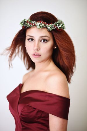 A woman in a burgundy off-shoulder dress wears a delicate flower crown against a white background.