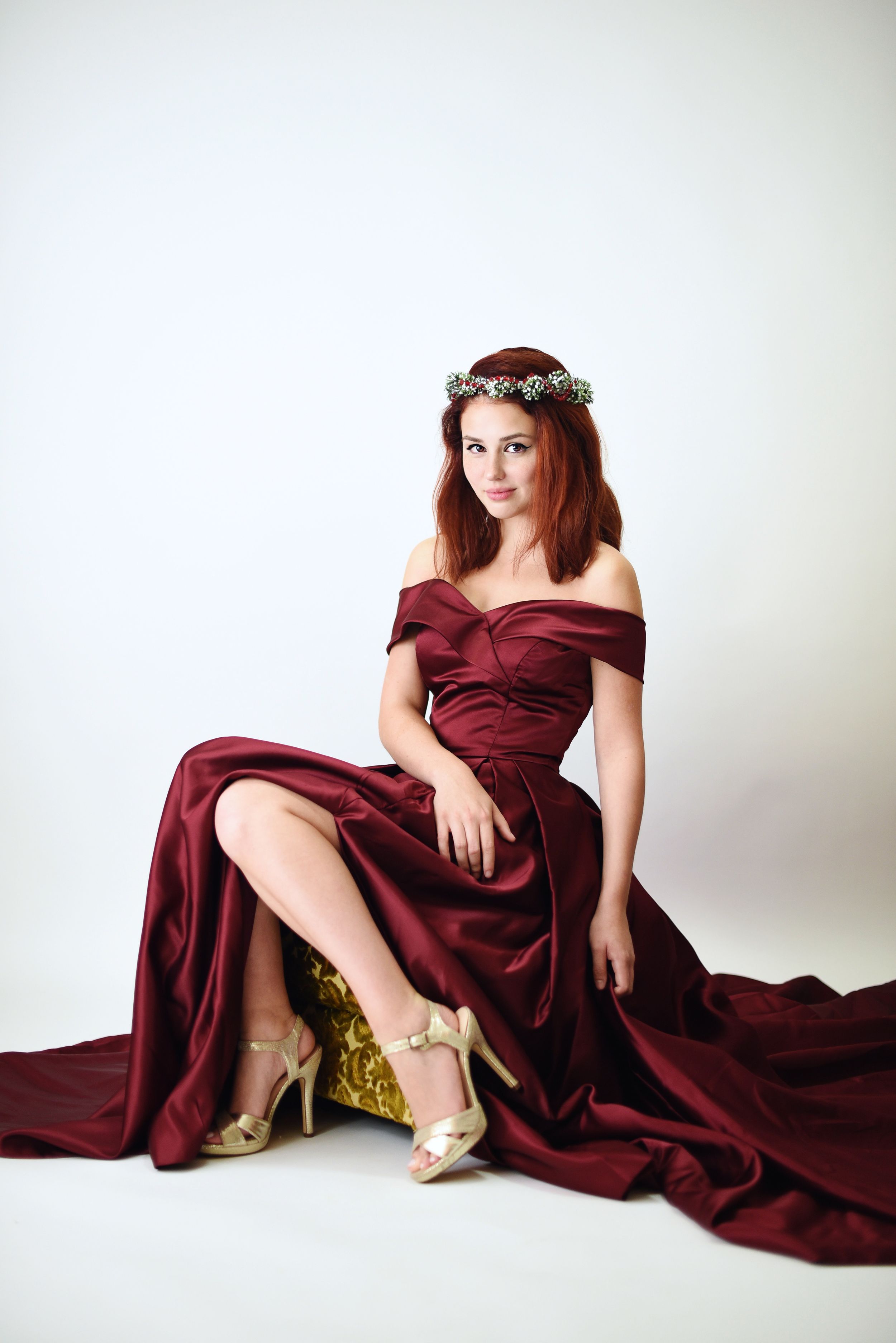 A woman in a burgundy evening gown and flower crown poses elegantly against a white backdrop.
