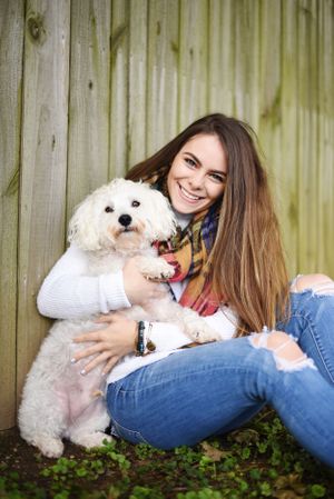 A cute white fluffy dog being hugged by someone wearing jeans and a white sweater against a wooden fence.