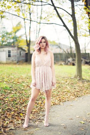 Portrait of person in blush lace dress standing on autumn pathway with fallen leaves.