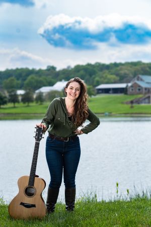 A musician poses with an acoustic guitar by a scenic lake with rolling hills and a barn in the background.