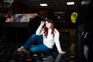 Woman in white sweater poses on colorful vinyl record floor in dark indoor setting.