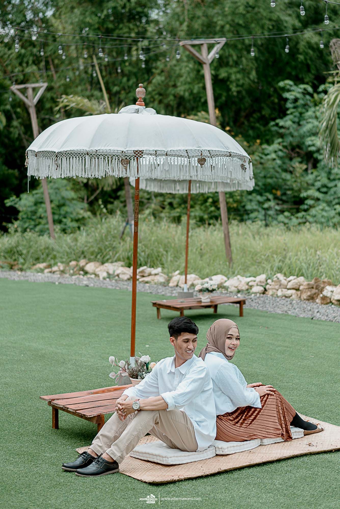 A stylish couple relaxes on a picnic blanket under a white fringed umbrella in a garden setting.