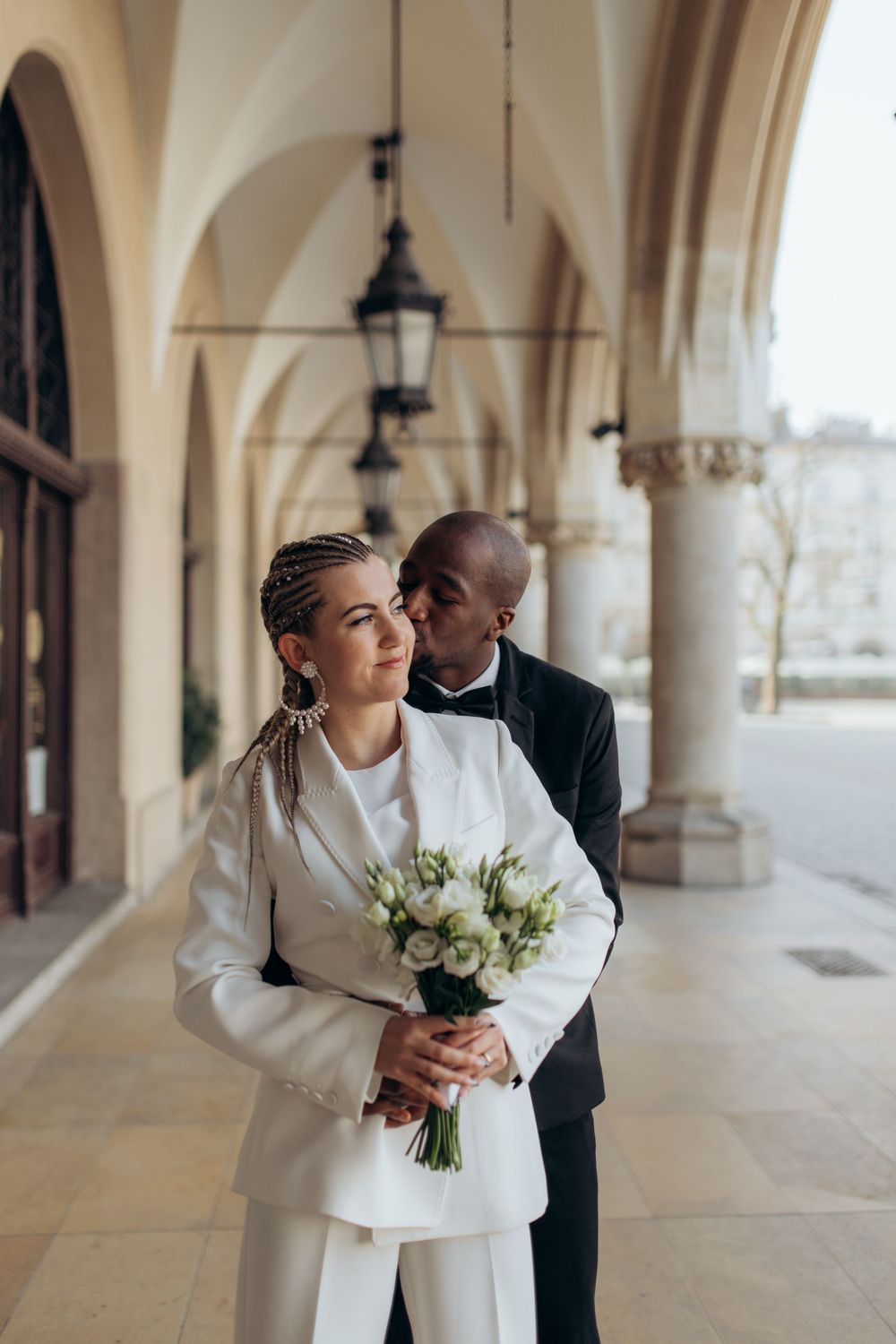 Couple in formal attire embracing under arched colonnade with white floral bouquet.
