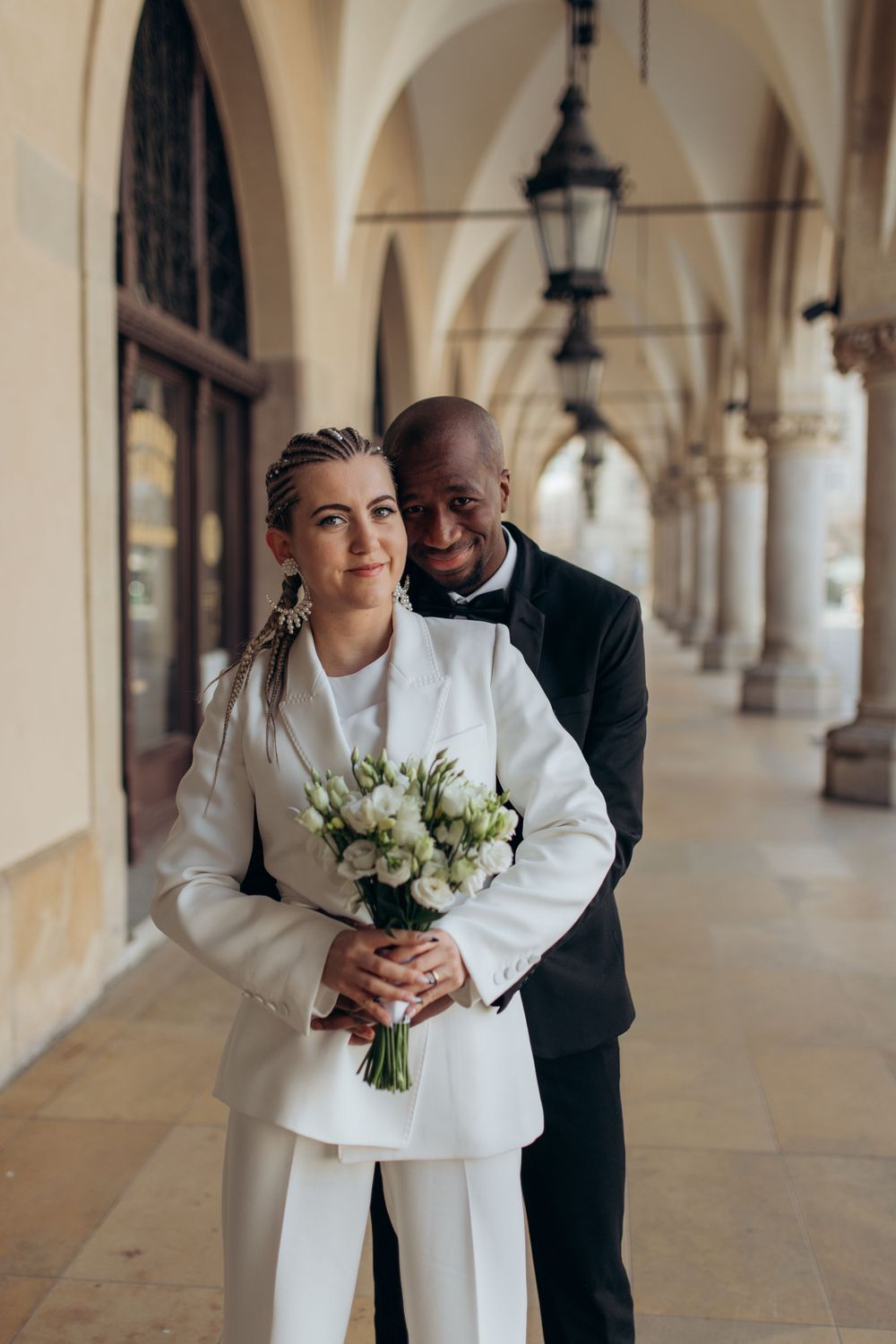 Wedding couple in white suit and tuxedo posing under ornate arched walkway.