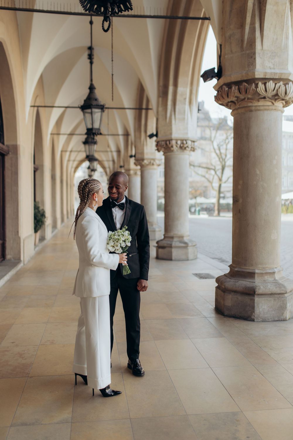 Elegant couple in formal attire standing in columned corridor with bouquet.