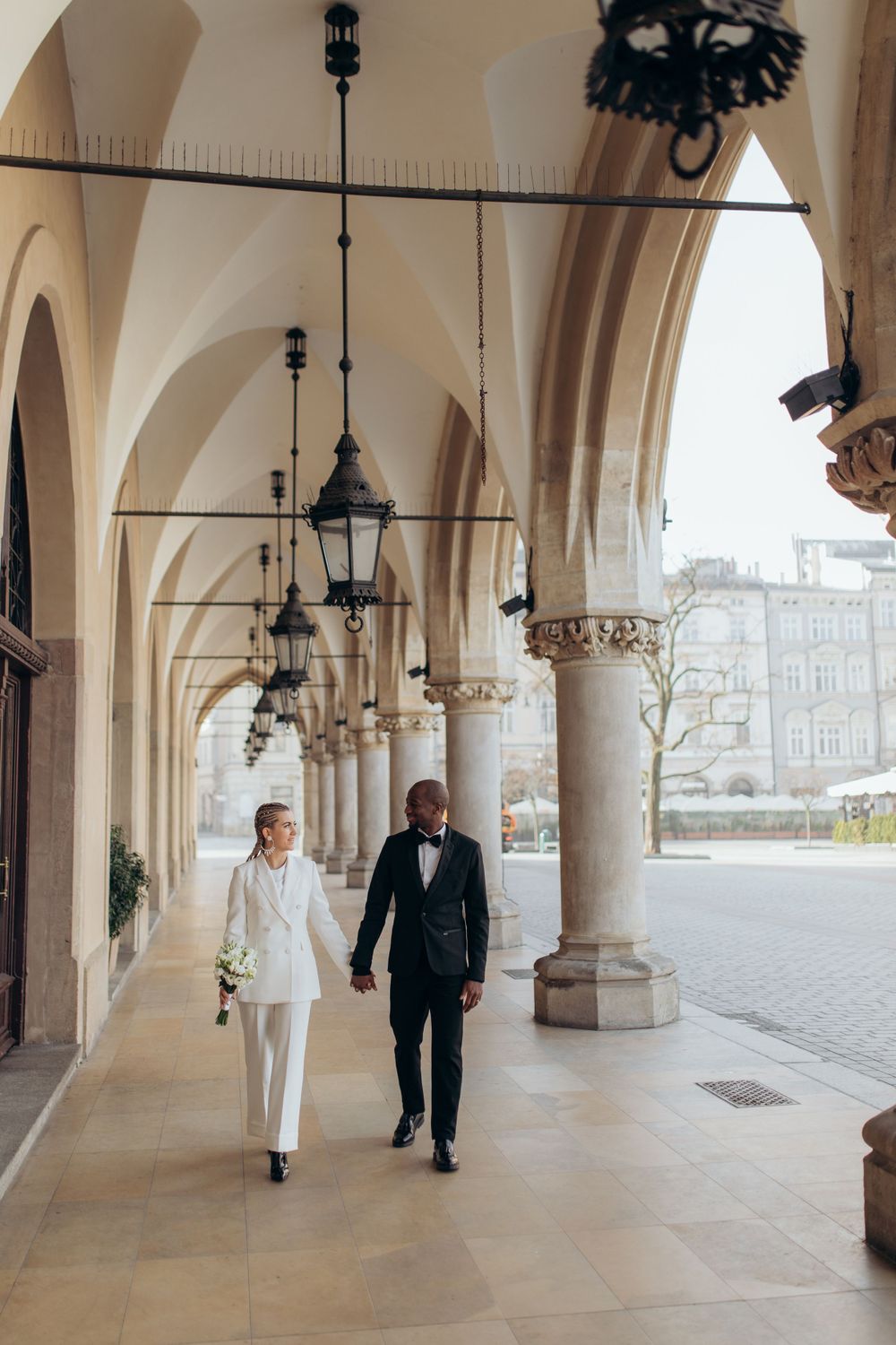 Couple walking together under elegant arched colonnade with hanging lanterns and ornate architectural details.