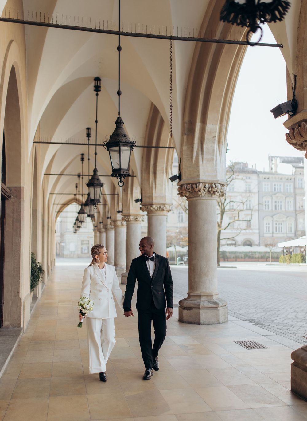 Elegant couple strolling through historic colonnade with vintage lanterns and stone columns.