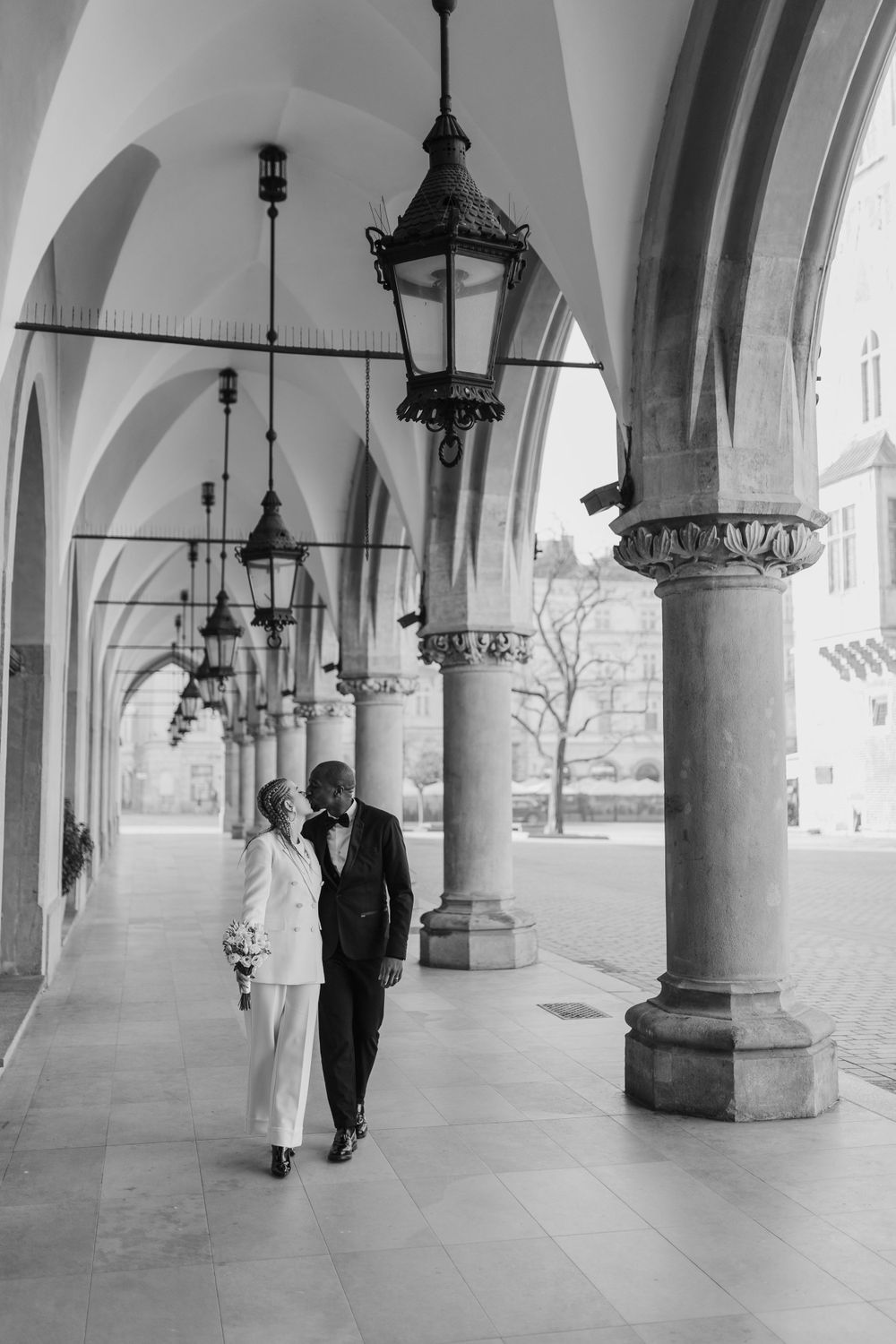 Black and white photo of couple walking along ornate covered walkway with gothic architecture.