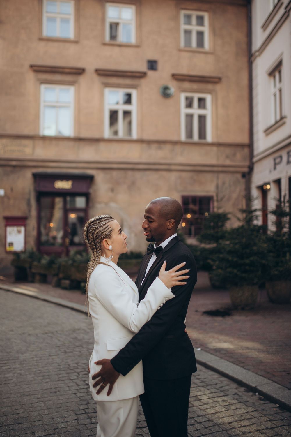 A romantic couple in formal attire embraces on a cobblestone street in front of a historic European building.