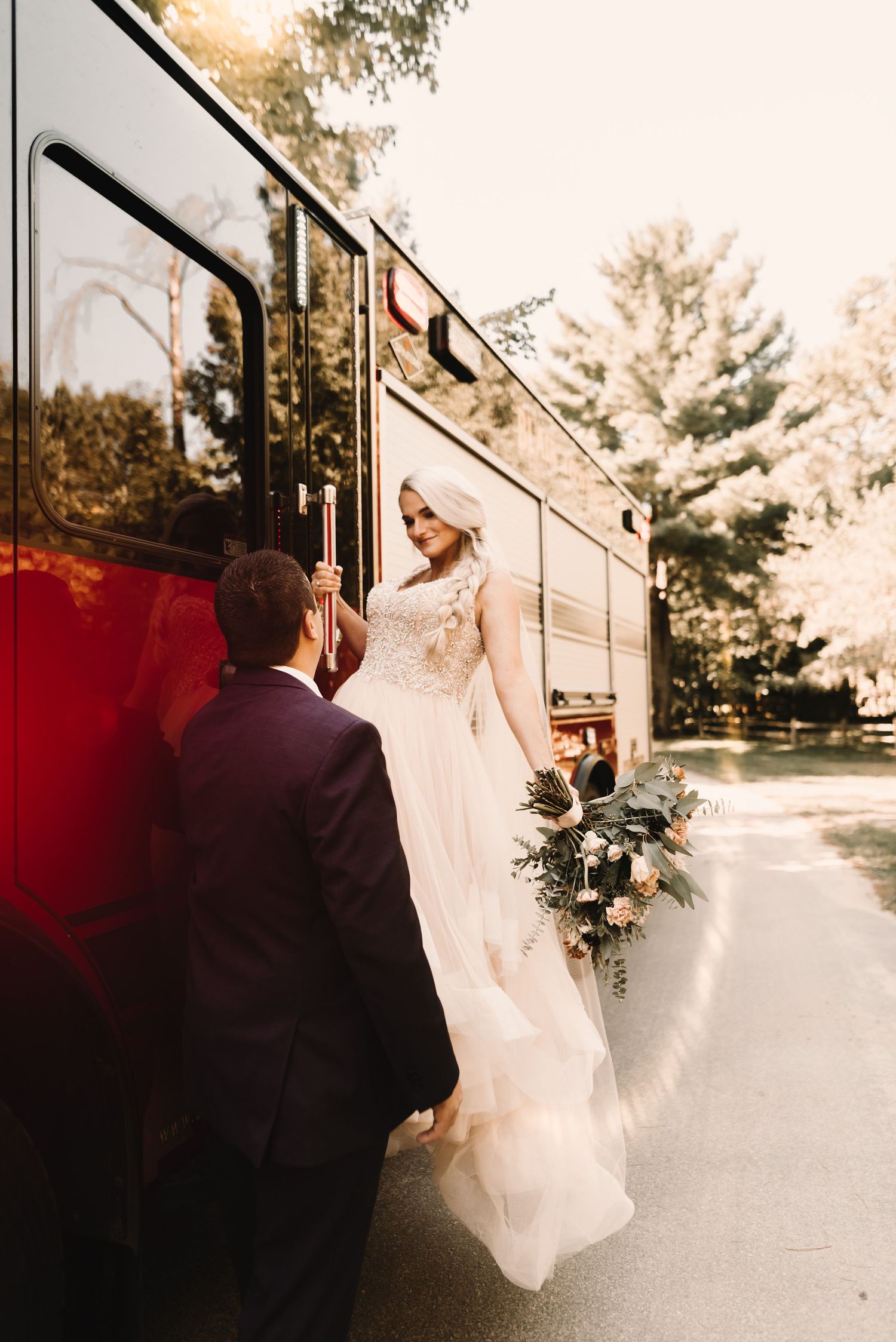 Bride in flowing white dress stepping onto vintage red bus while groom assists.