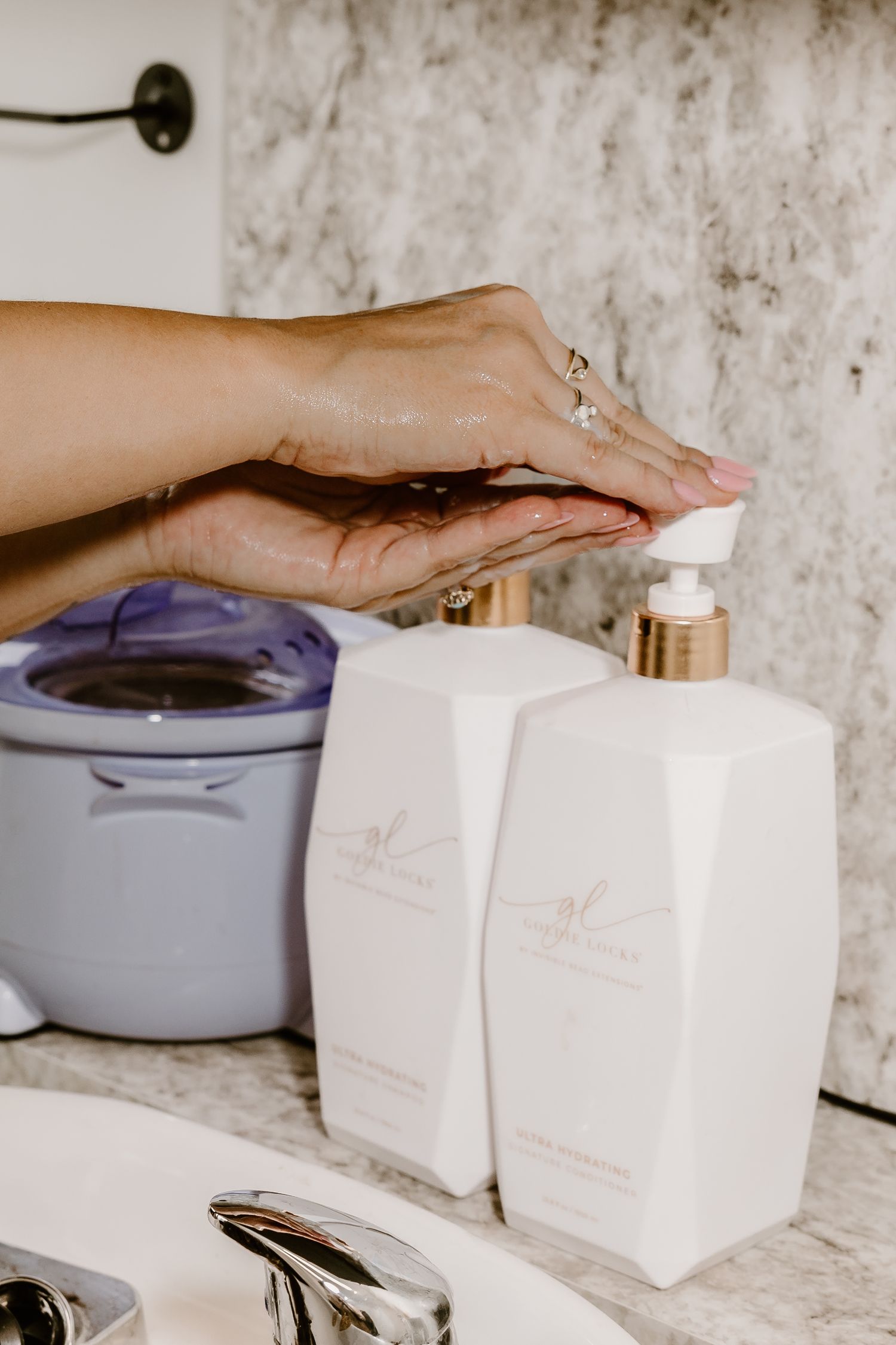 Hands applying beauty products from white bottles on marble counter with purple device.