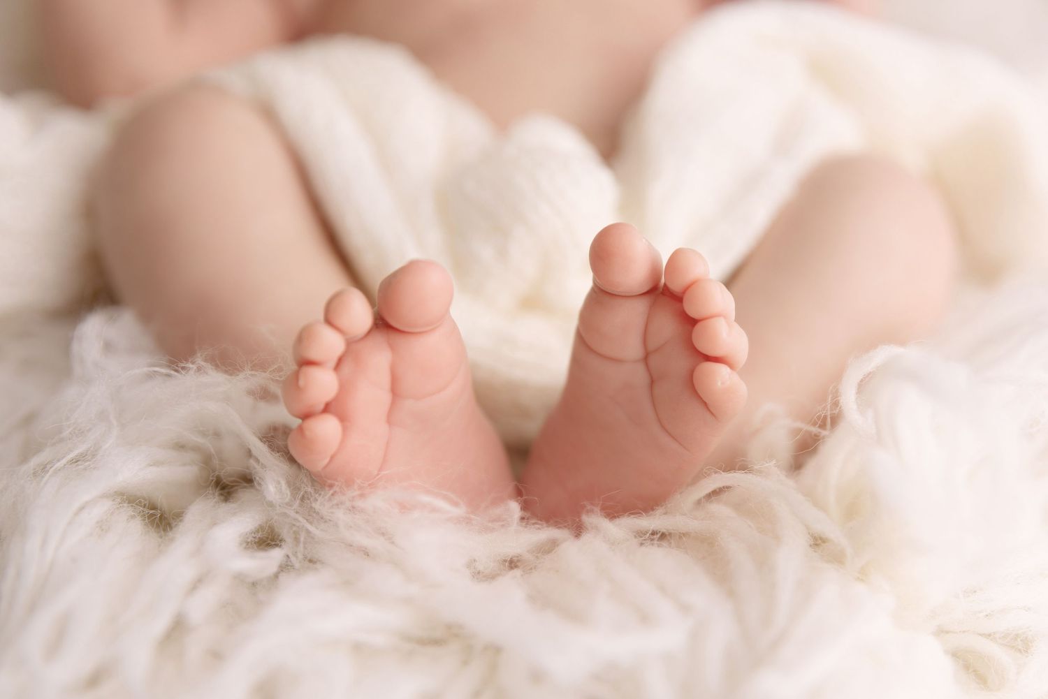 Tiny newborn baby feet resting on soft white fur blanket in gentle natural lighting.