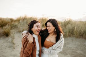 Two friends sharing laughter together on beach with sand dunes behind them.