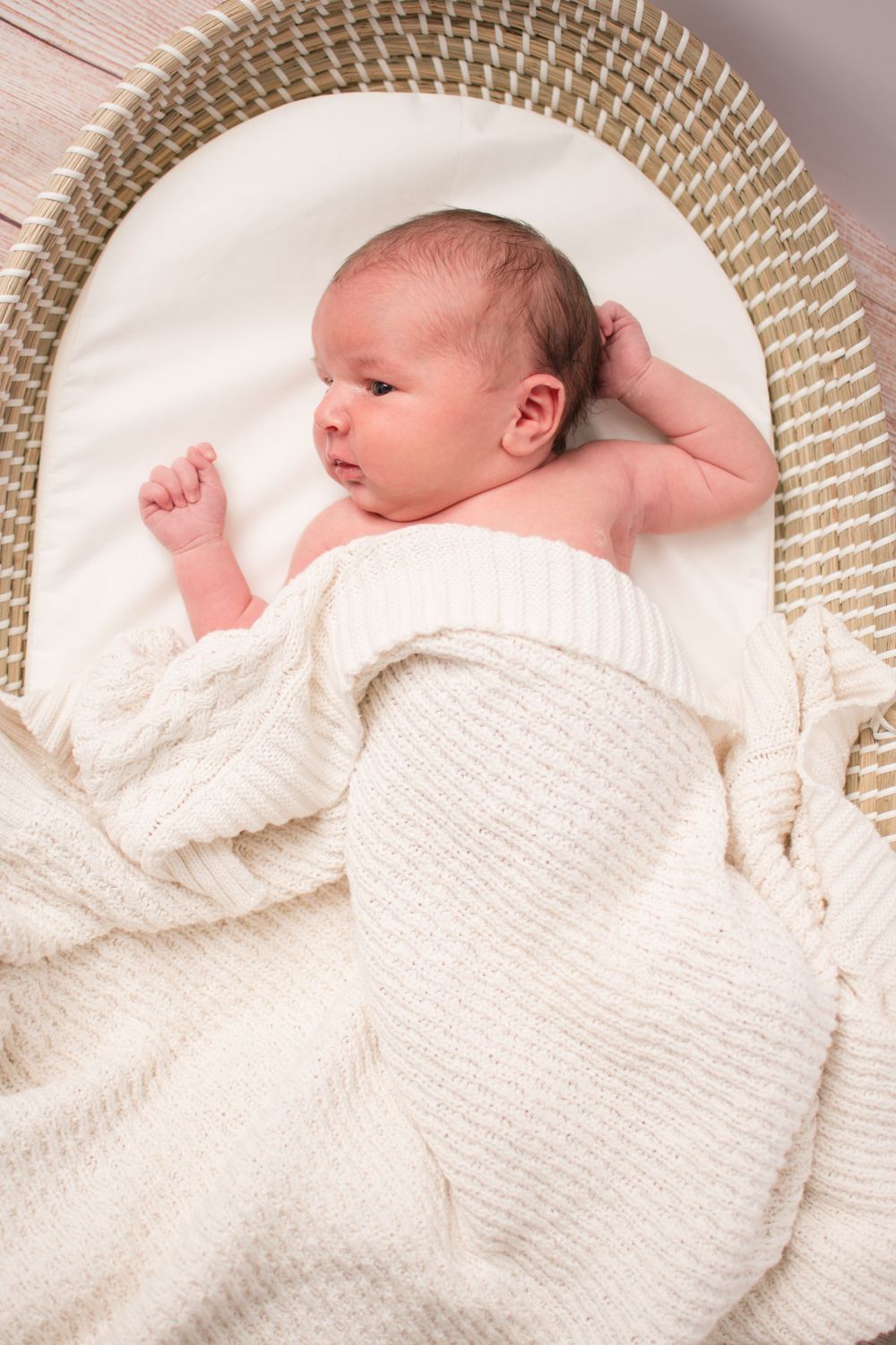 little one in Moses basket as seen from above. she is laying under a cream knit blanket and touching her hair.