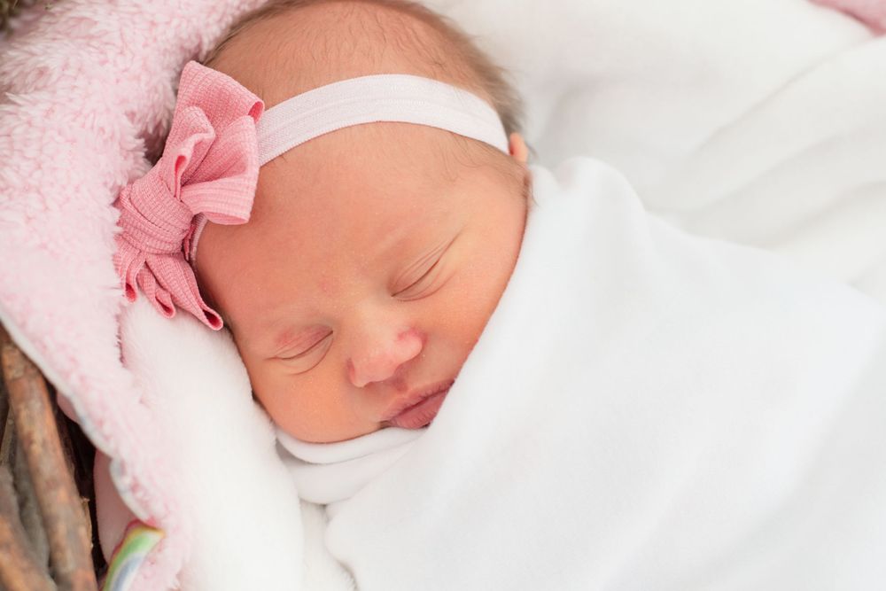 swaddled and sleeping baby. white swaddle with pink bow. she is laying in a basket with blankets