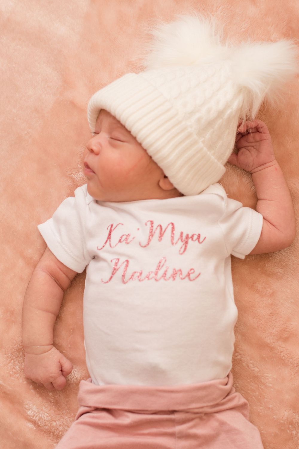 view from above of a little one in pink pants and white onesie with a cream hat and fuzzy ball top. She is sleeping