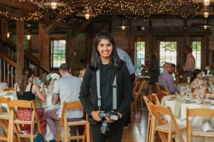 A photographer with camera gear stands in a rustic venue decorated with string lights.
