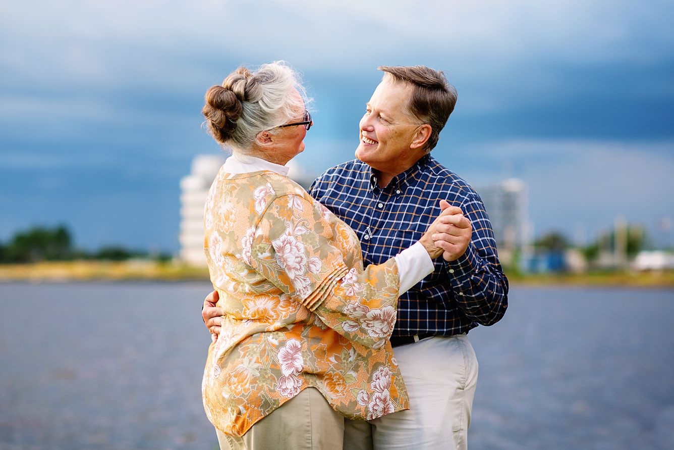 Happy senior couple dancing together outdoors near a lake under a dramatic cloudy sky.