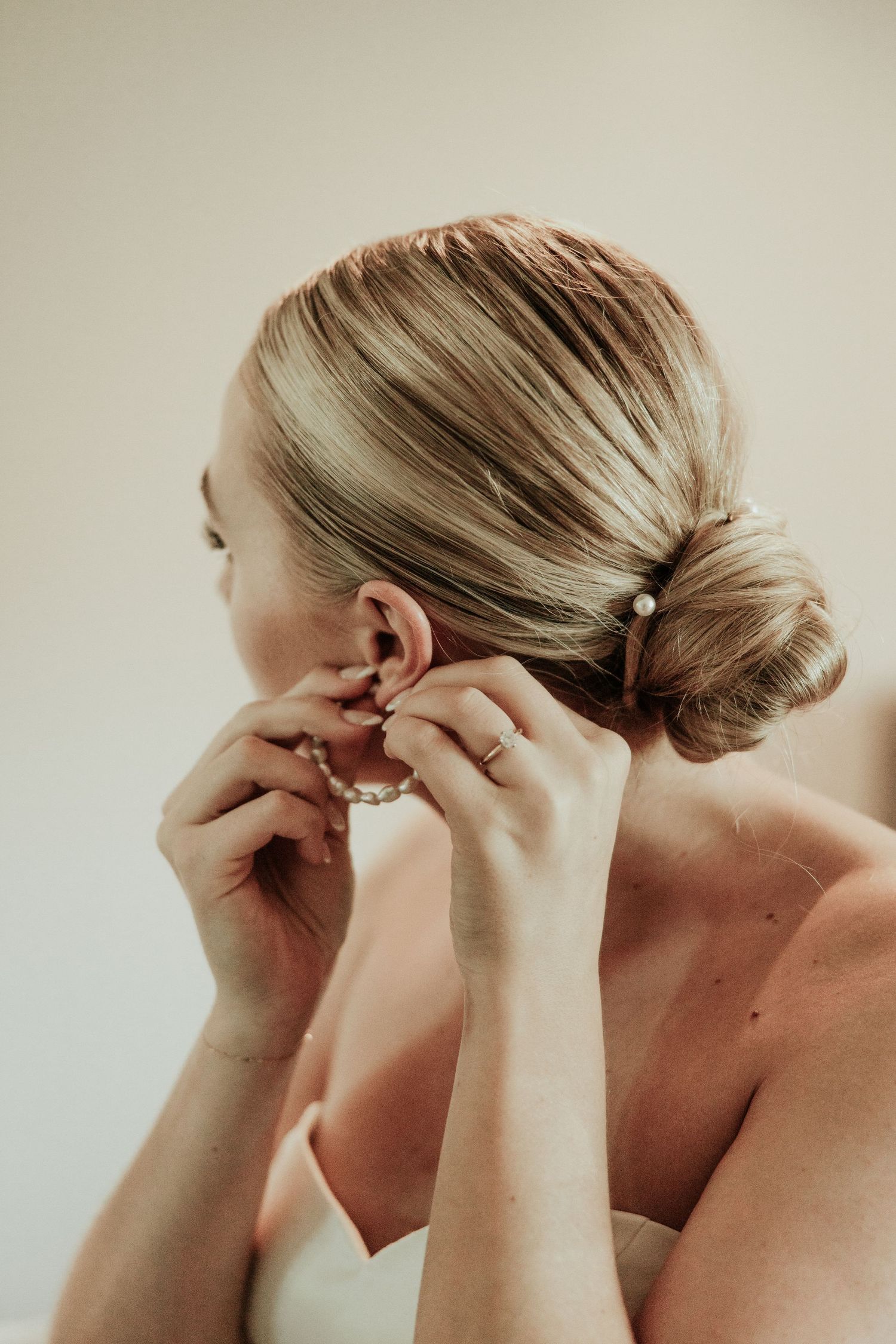 Woman with blonde hair in a low bun puts on earrings against neutral background.