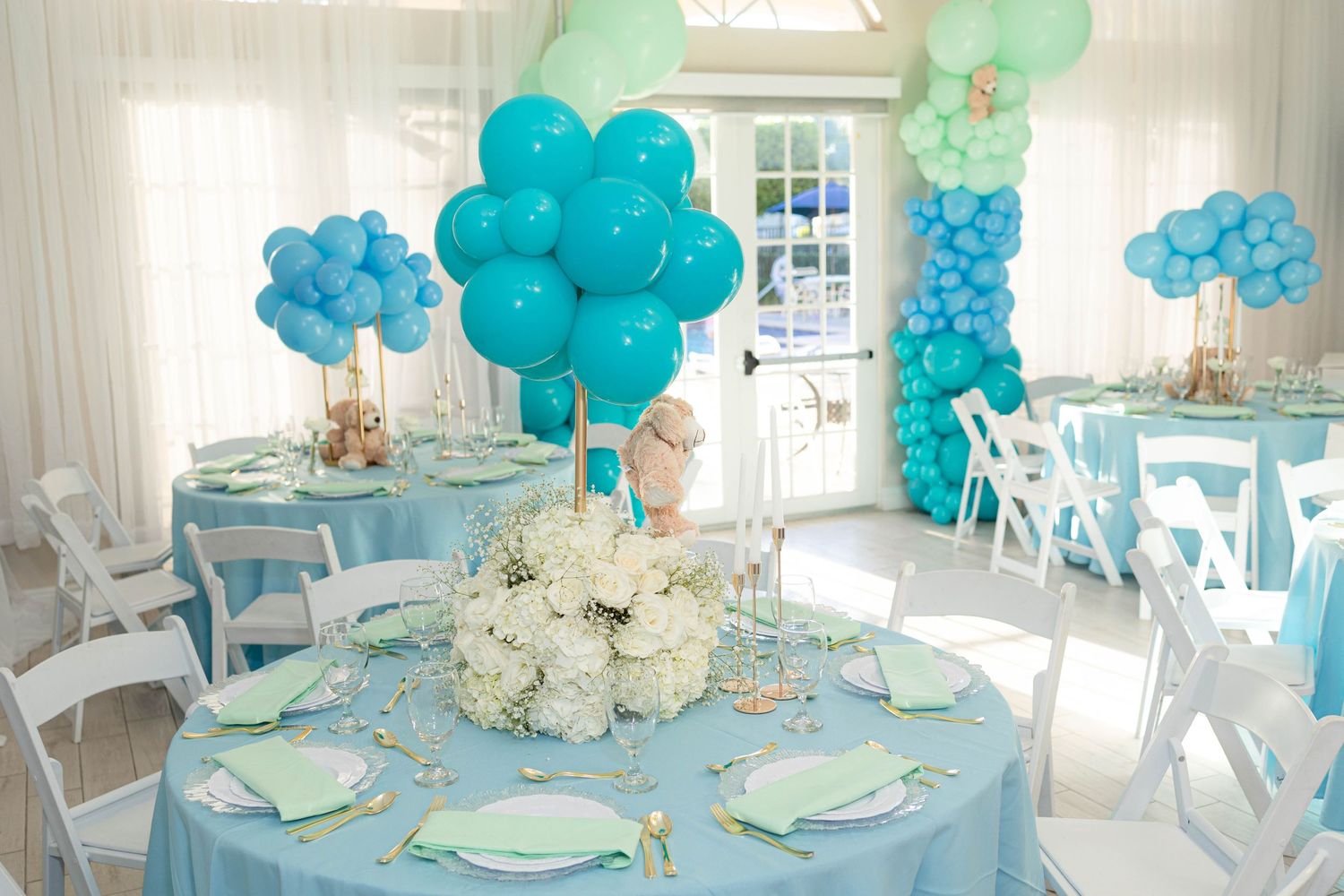 Blue and mint baby shower table setting with balloon centerpieces and white floral arrangements.