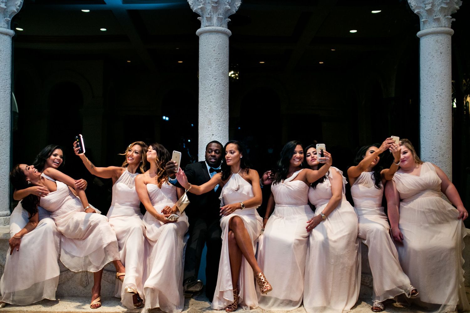 A group of bridesmaids in light pink dresses pose together in an elegant columned venue.