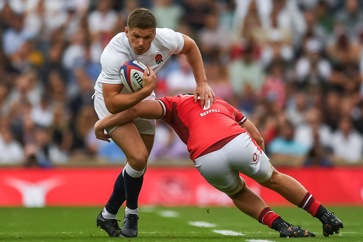 Rugby player in white jersey gets tackled by opponent in red during intense match action.