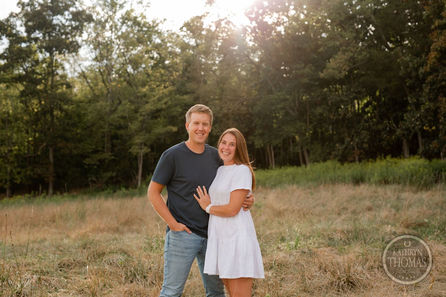 A romantic couple embraces in a golden field with sunlight streaming through trees in the background.