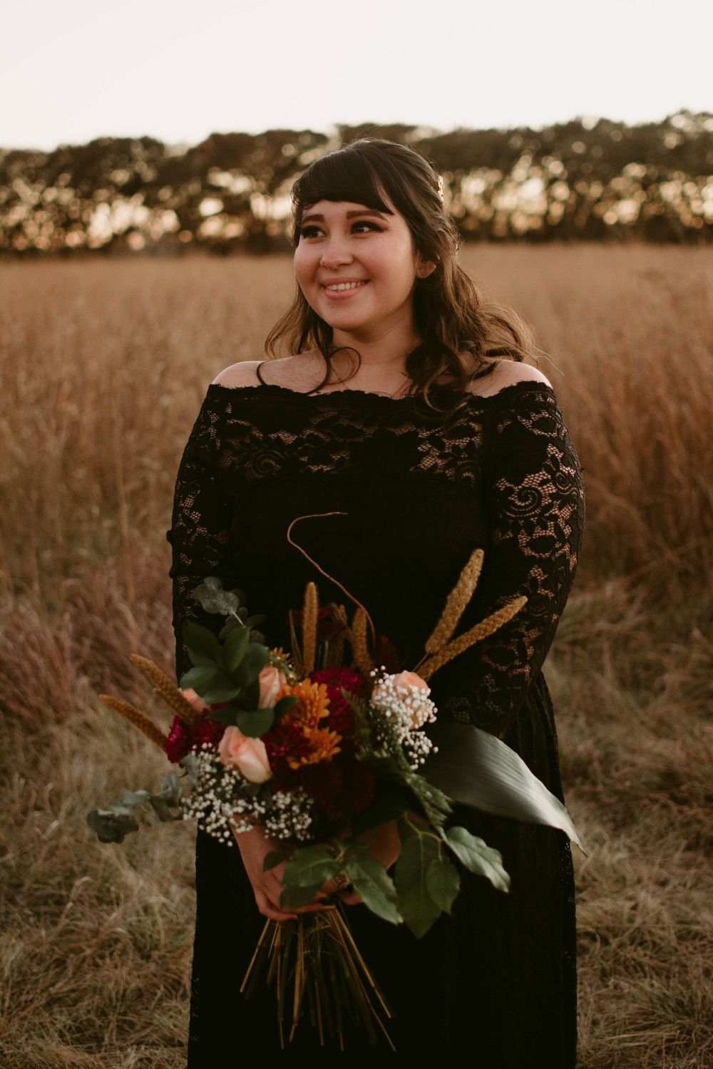 A person in black lace carries a vibrant bouquet of flowers in a rustic field setting.