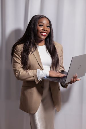 Business professional in tan blazer working on laptop against neutral background.