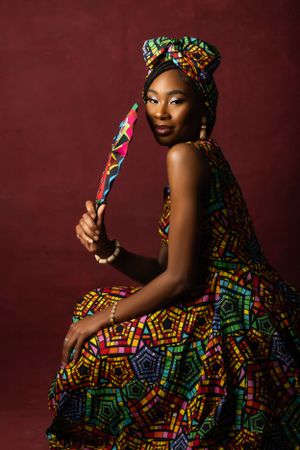 Woman in vibrant African print dress and headwrap holds decorative braid against burgundy background.