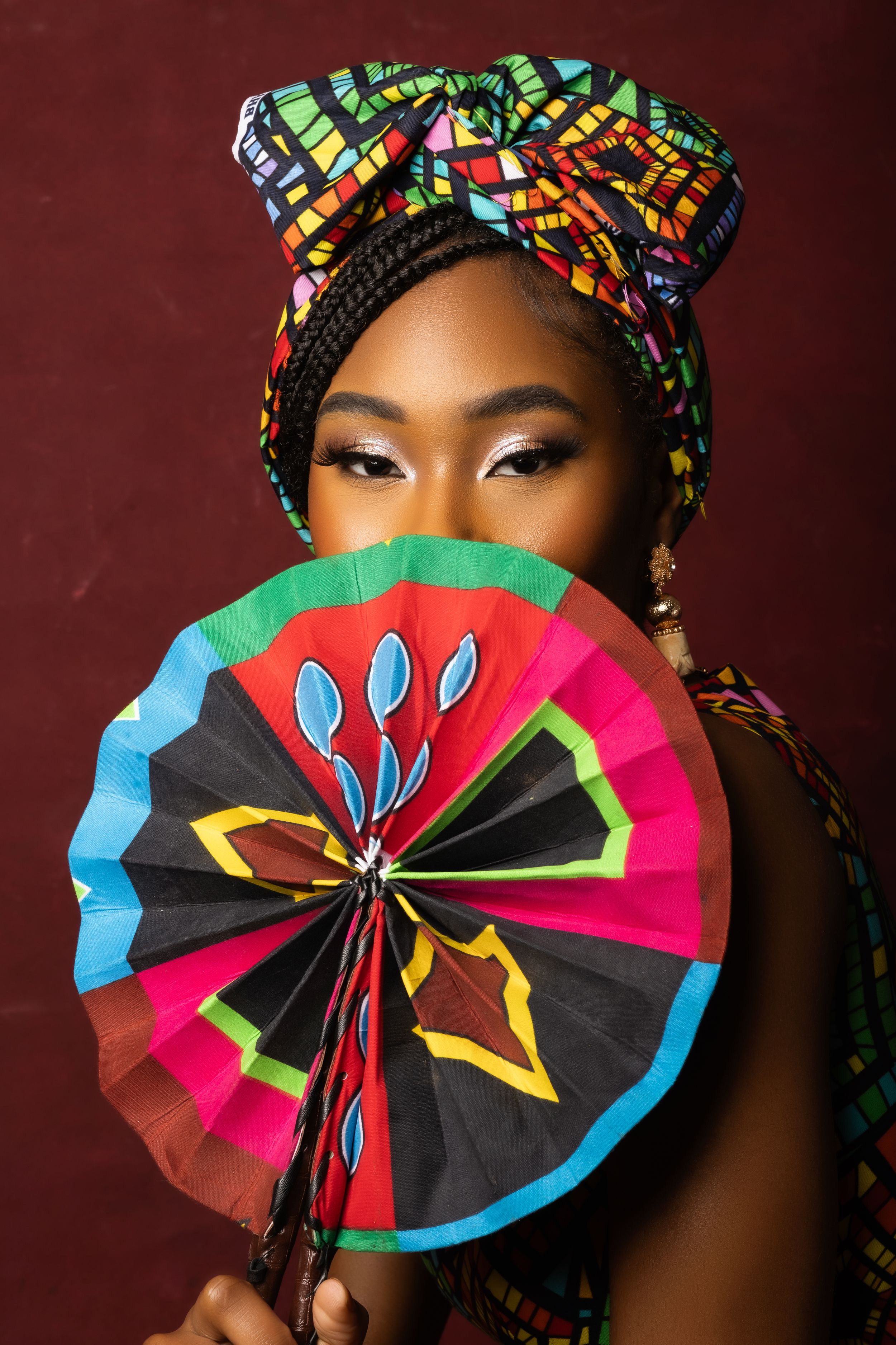 Woman in traditional headwrap holds colorful decorative fan against burgundy backdrop.