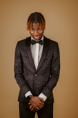 Portrait of person in patterned black tuxedo and bowtie against warm brown background.