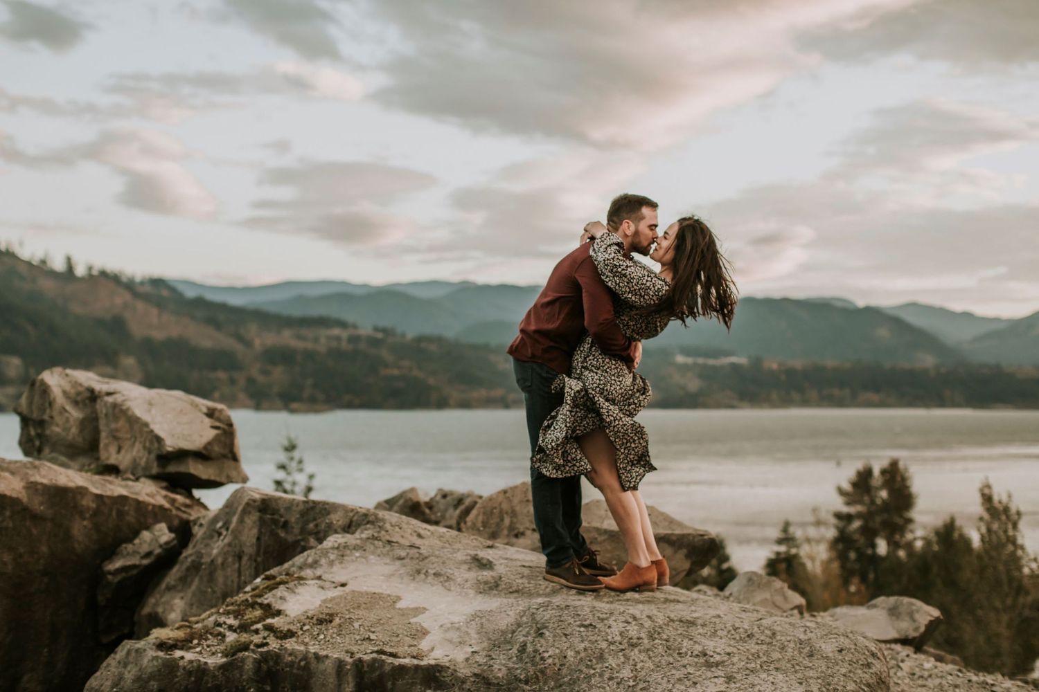 Girl and boy hugging and kissing while standing on a rocky formation with the Columbia River Gorge in the distance