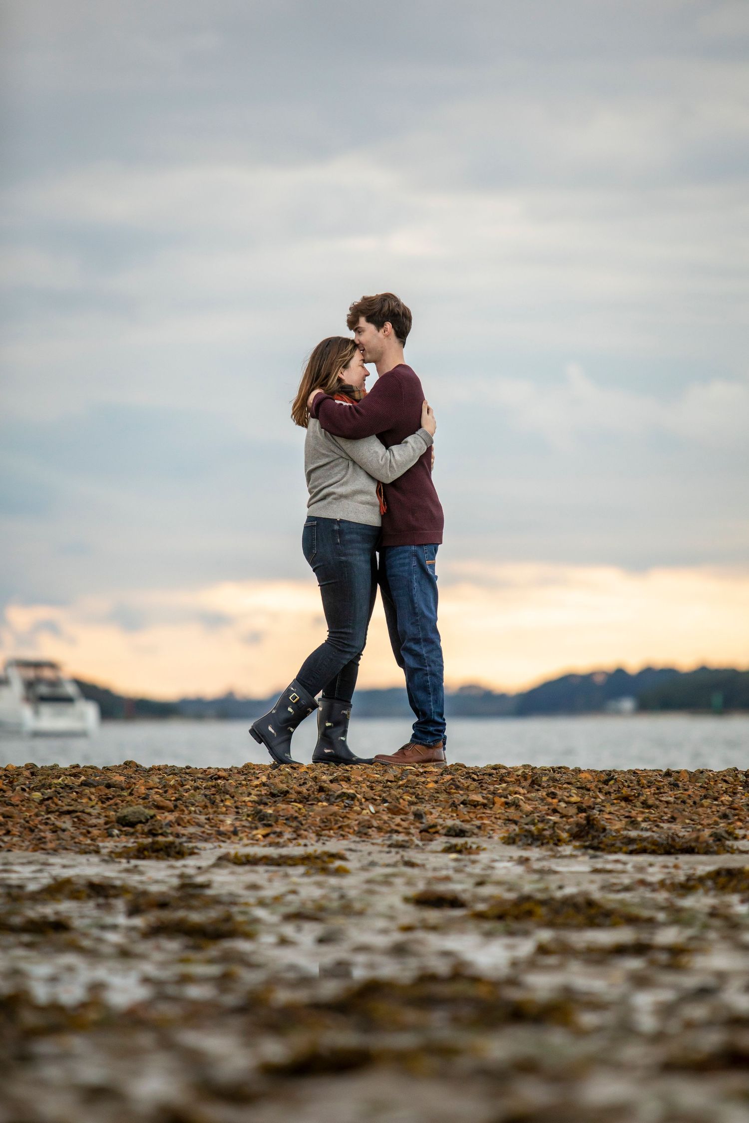 Couple embracing on a rocky beach at sunset with water and boats in background.