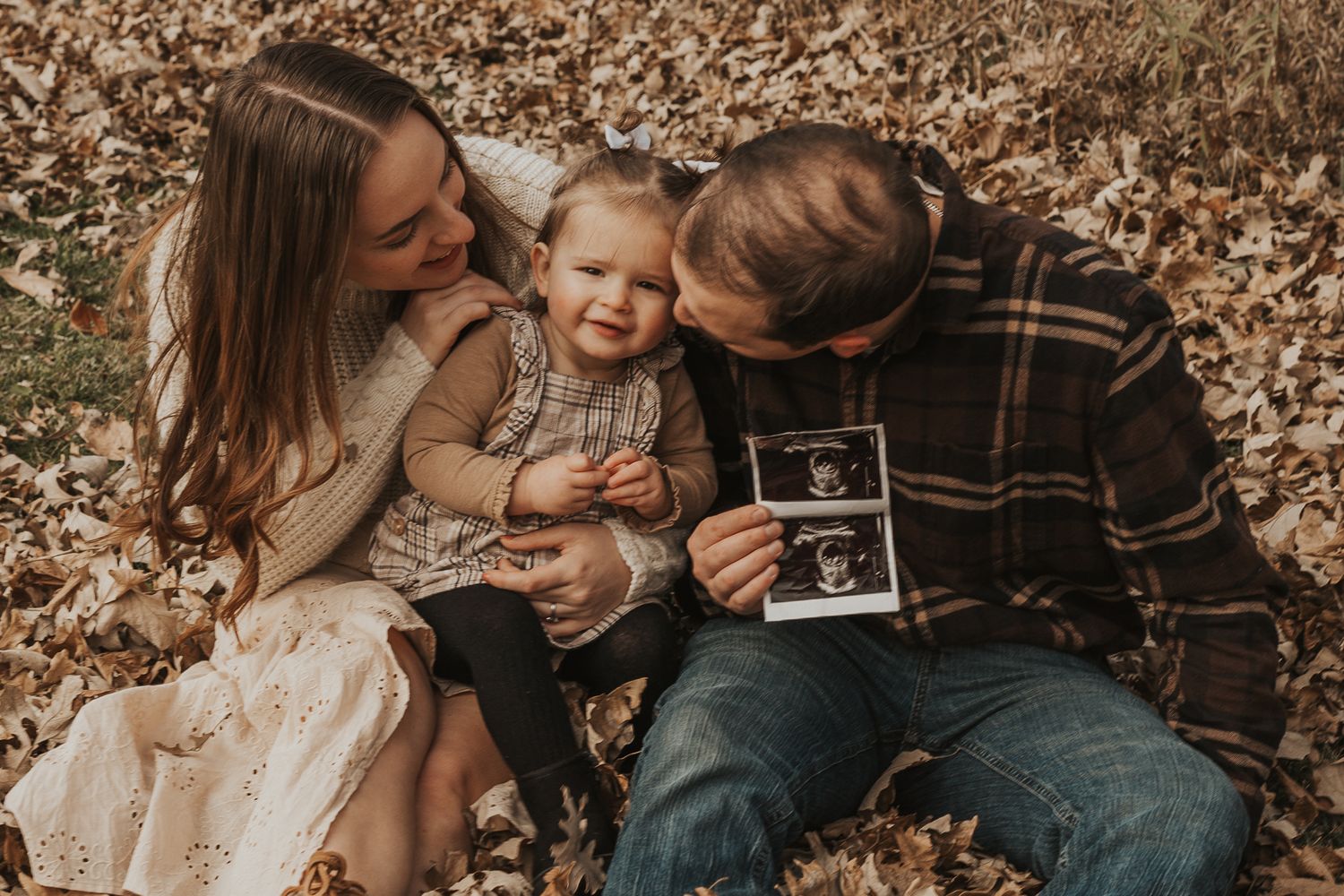 Family shares intimate moment on autumn leaves while announcing pregnancy taken by Michigan maternity photographer.