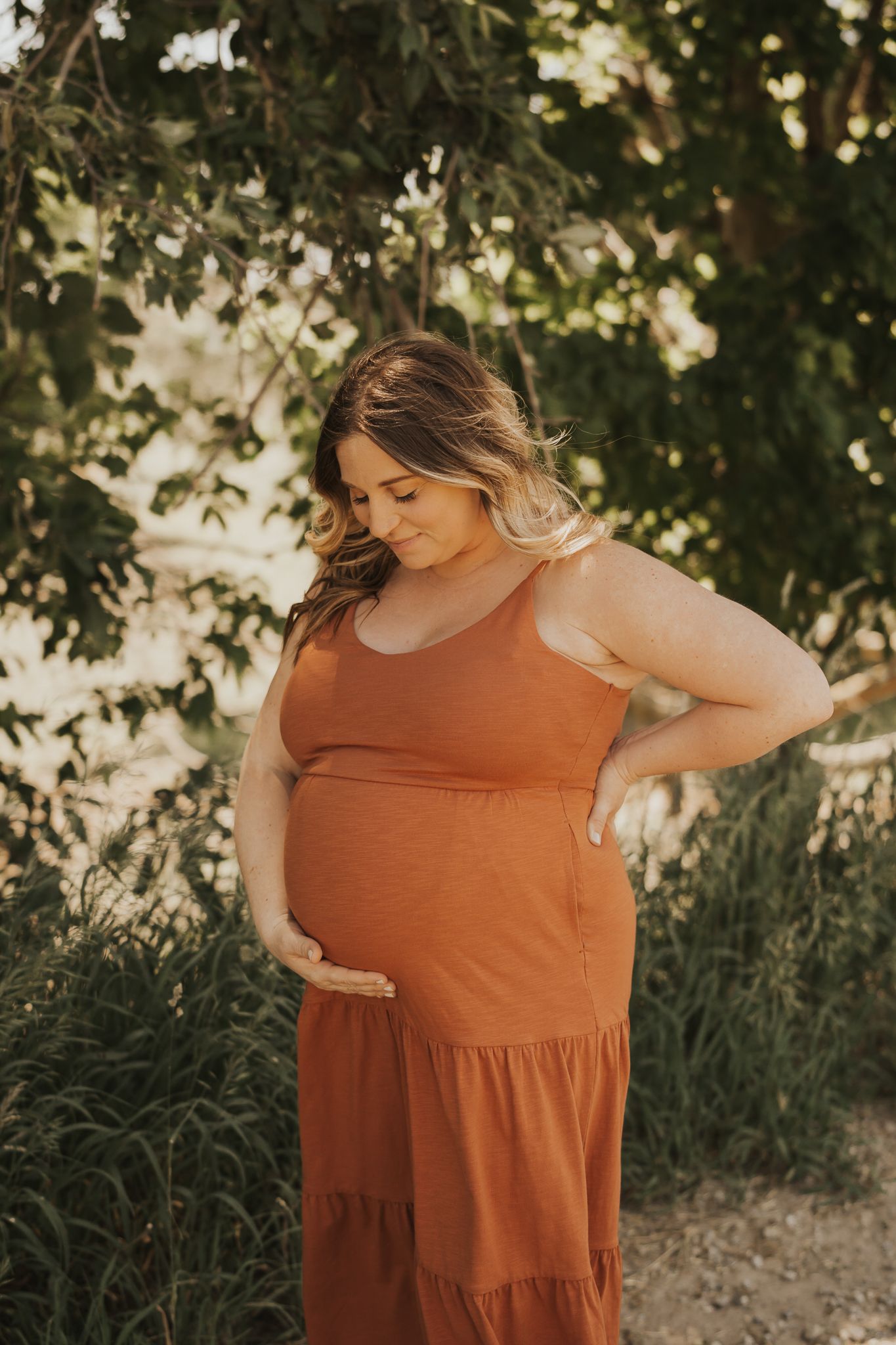 Expectant mother in rust-colored dress poses under leafy trees during maternity shoot.