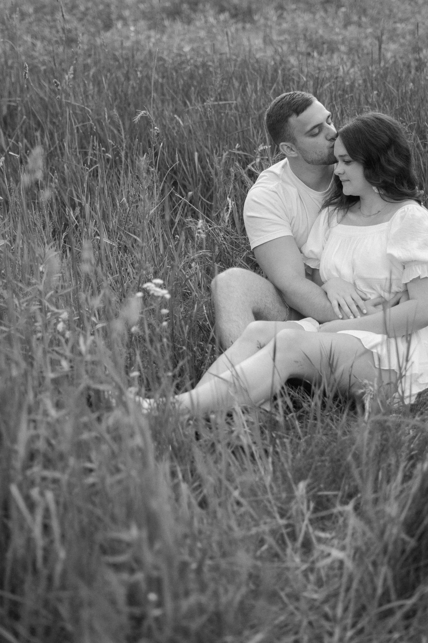 Romantic couple sitting in tall grass field during sunset in black and white.
