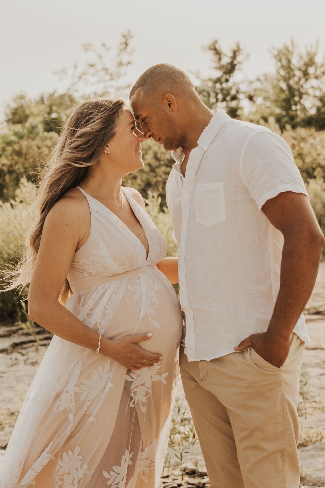 Expecting couple in white outfits share intimate moment during sunset photoshoot.